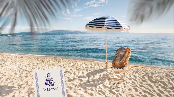 Slunečná pláž v Řecku s modrobílým slunečníkem, pod kterým na lehátku odpočívá bochník chleba – vizuál ke kurzu pečení s Mari Peče v Athénách v říjnu 2025.